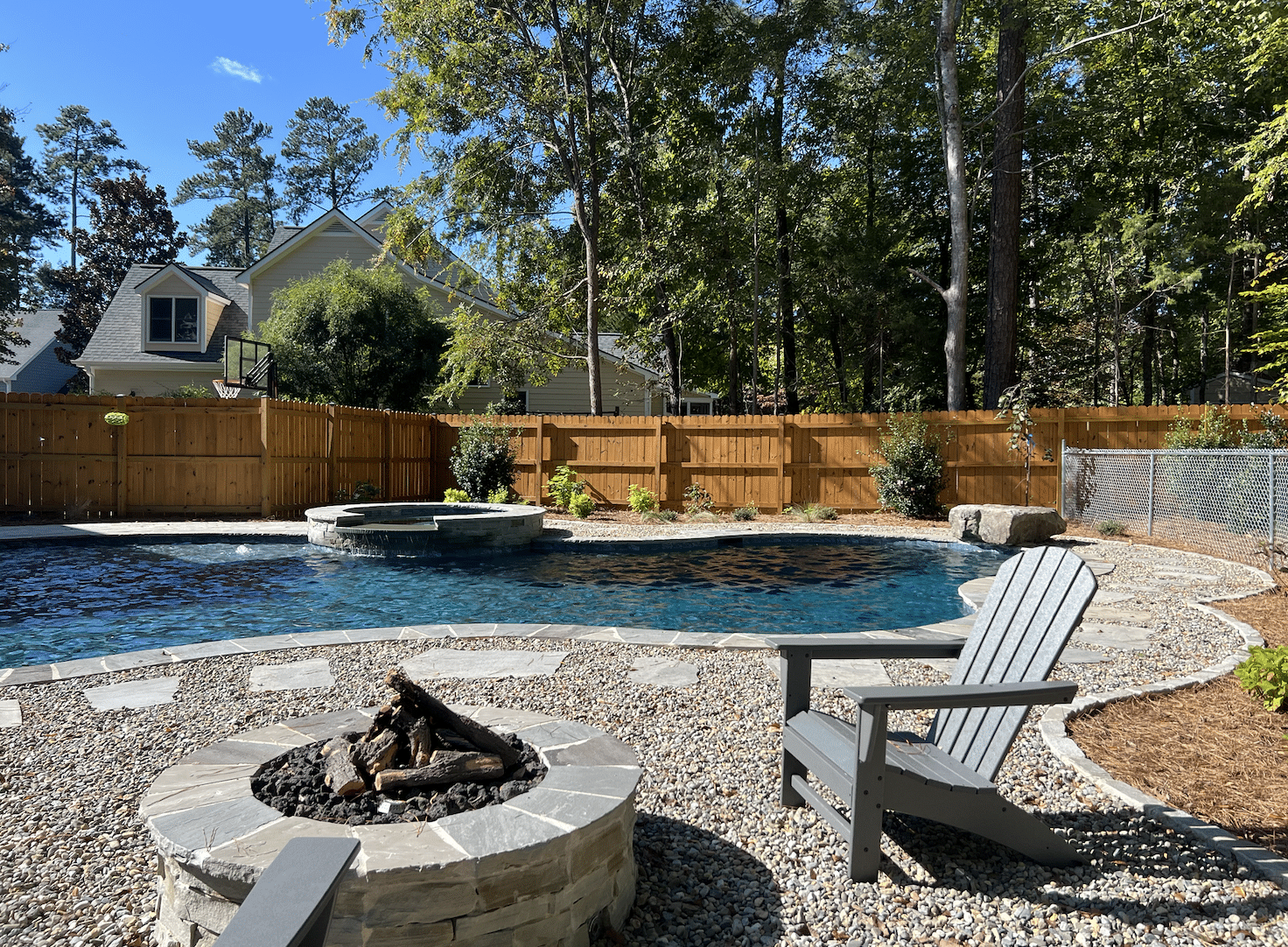 A backyard pool with integrated fire pit, stone patio, wooden fencing, and Adirondack chairs surrounded by mature trees and a residential home.